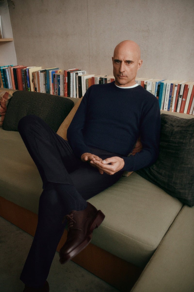 Mark Strong sits on a sofa in a dark crewneck sweater and dark trousers.