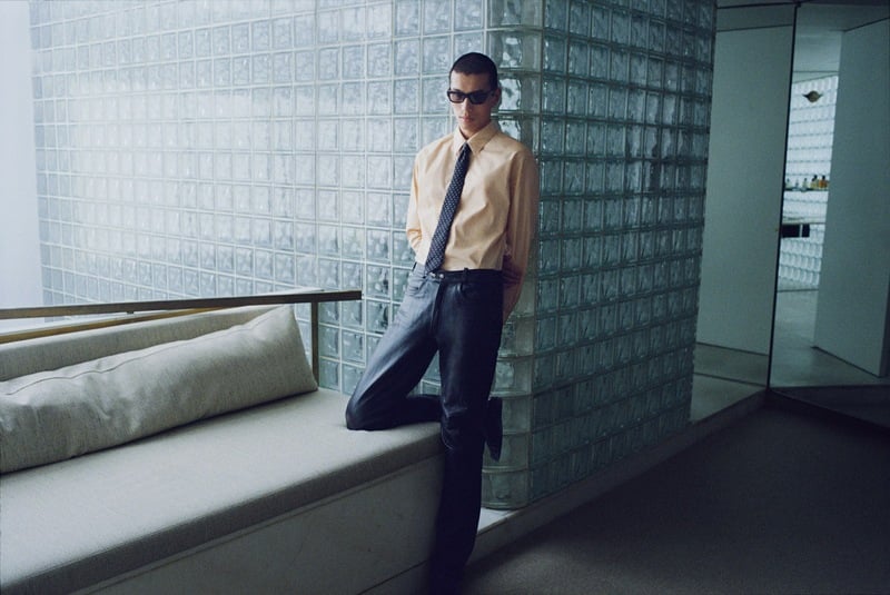 Yuyang Cai wears sunglasses, a beige short-sleeve shirt, dotted tie, and dark trousers beside a glass-block wall.
