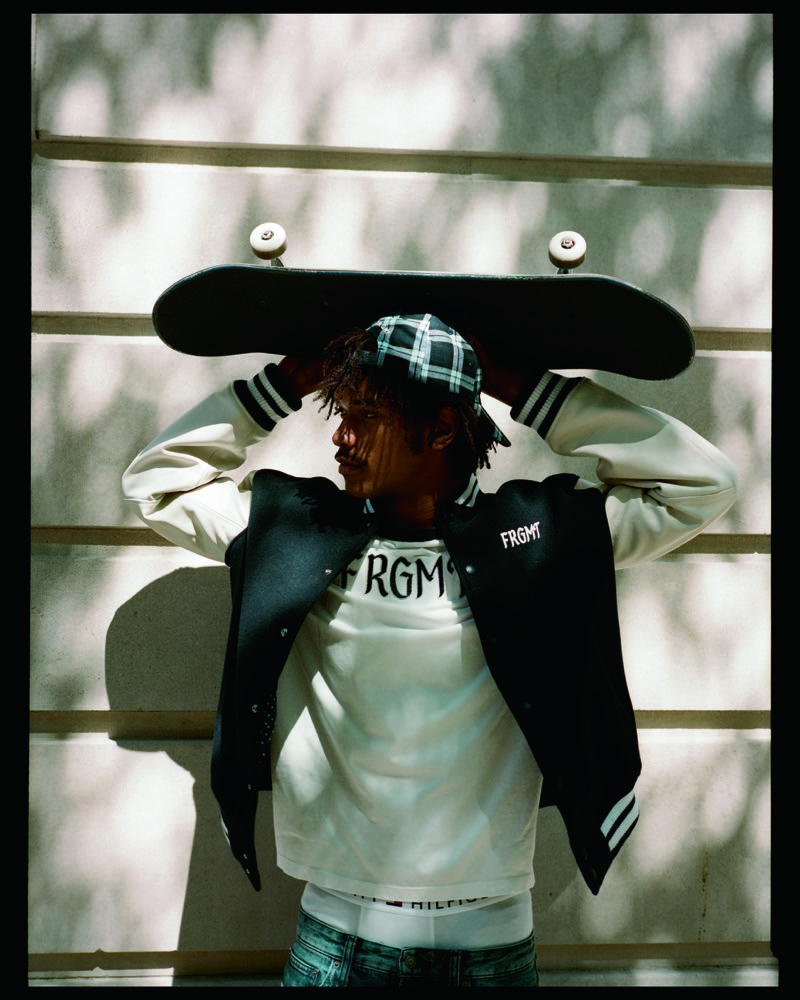Nile Winchester holds a skateboard above his head in a varsity jacket, graphic T-shirt, and plaid cap.