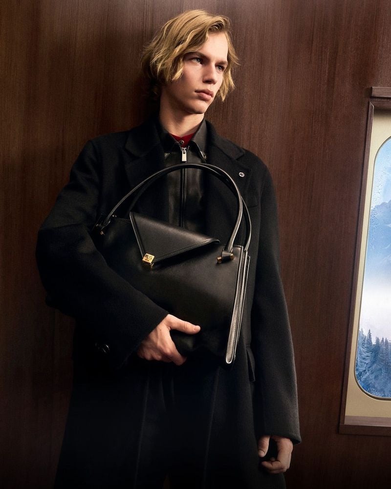 Max Stenzel holds a black handbag against his black coat inside a wood-paneled train carriage.