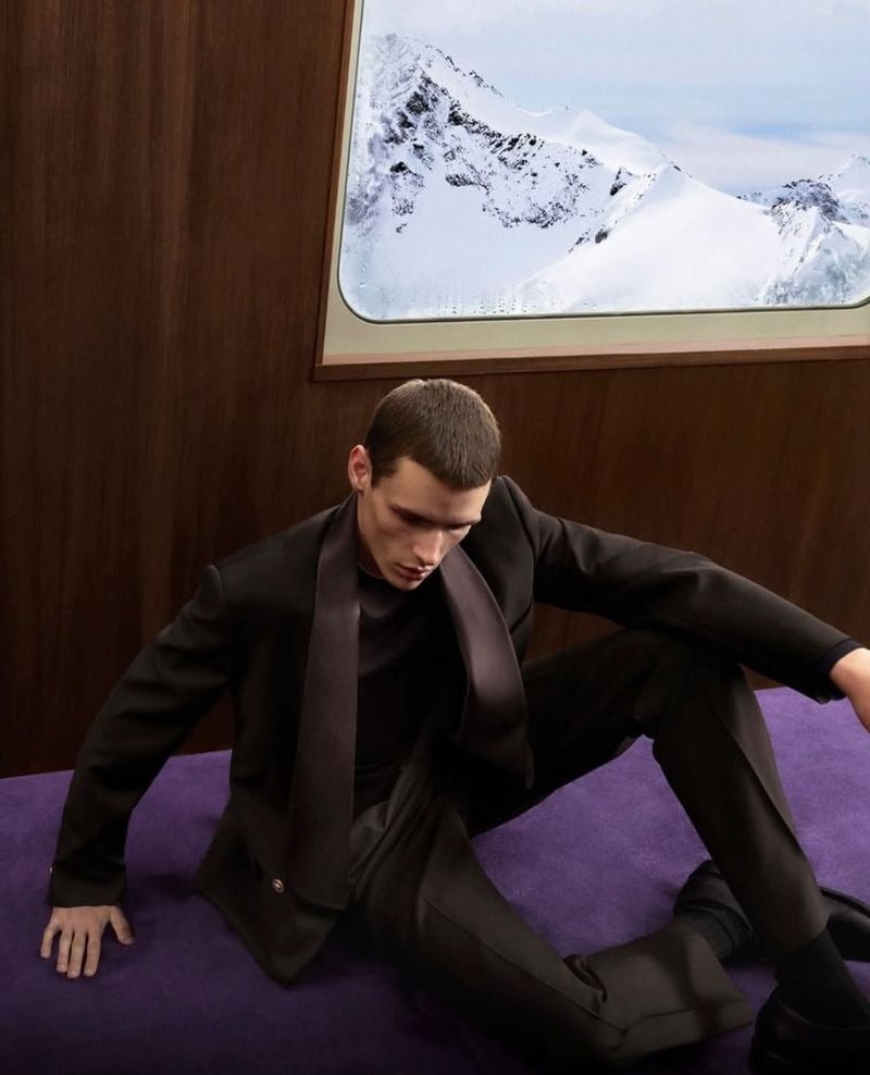 Paolo Grebic lounges on a lavender train seat wearing a suit and long scarf.