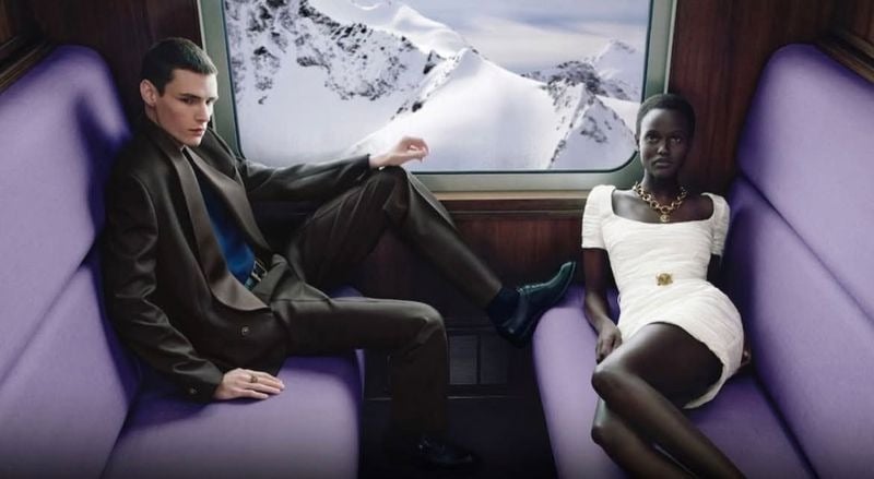 Paolo Grebic wears a suit and scarf opposite Yar Aguer in a white dress and gold necklace, with snowy mountains outside.
