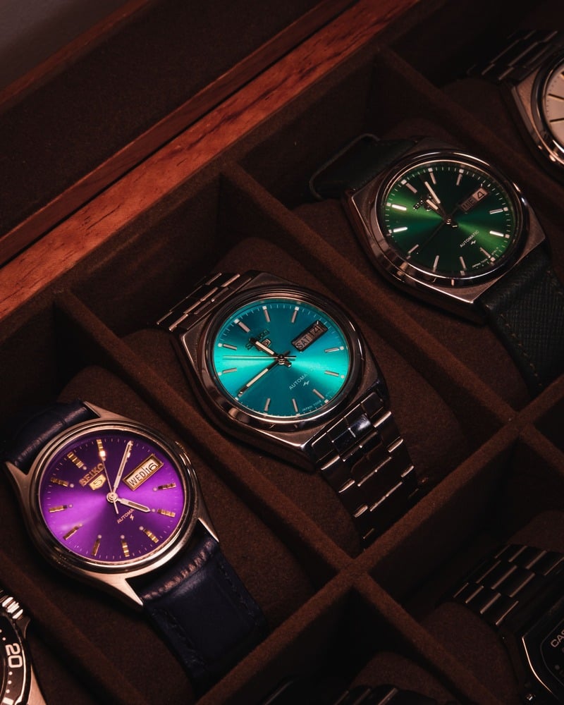 Watches with purple, turquoise and green dials in separate compartments of a wooden storage box.