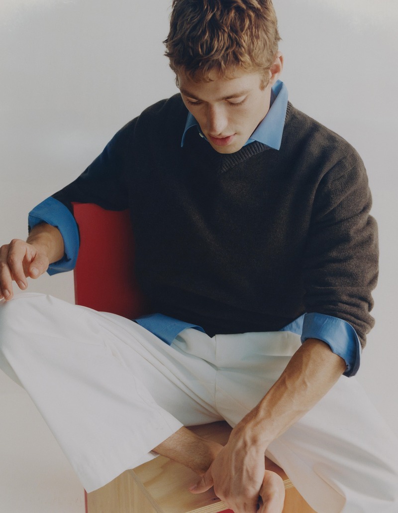 Valentin Humbroich looks down while seated in a dark sweater, blue shirt, and white trousers.