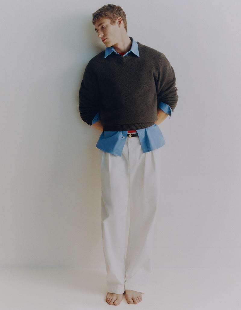 Valentin Humbroich stands barefoot in white trousers and a dark sweater with an exposed blue shirt hem.