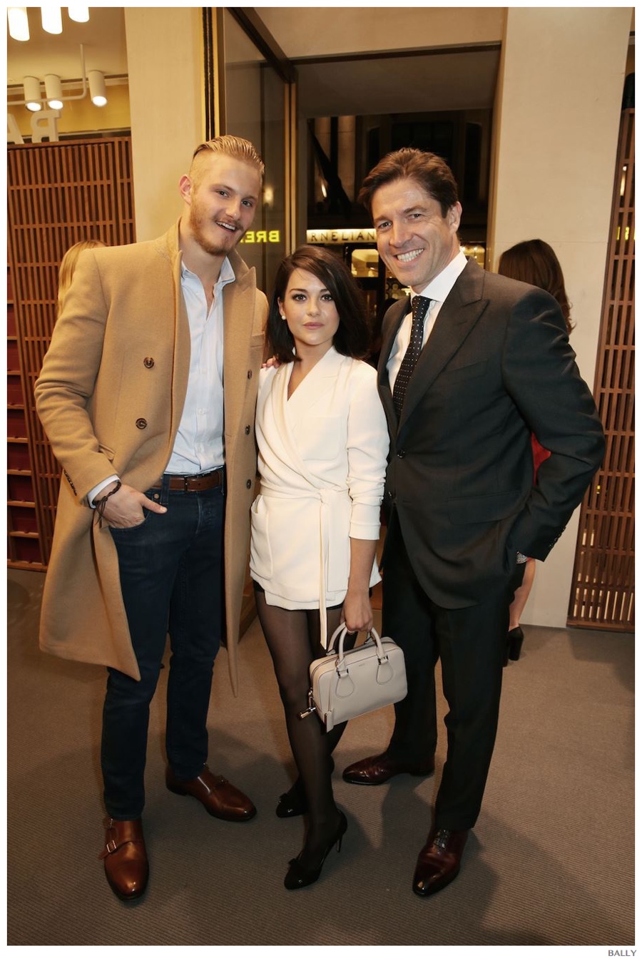 Dapper in a camel coat, actor Alexander Ludwig poses for a photo with actress Sarah Greene and Bally CEO Frédéric de Narp.