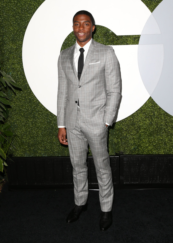 Chadwick Boseman wears Gucci light grey plaid notch lapel two-button Dylan suit with a white shirt, black tie and black leather lace ups.