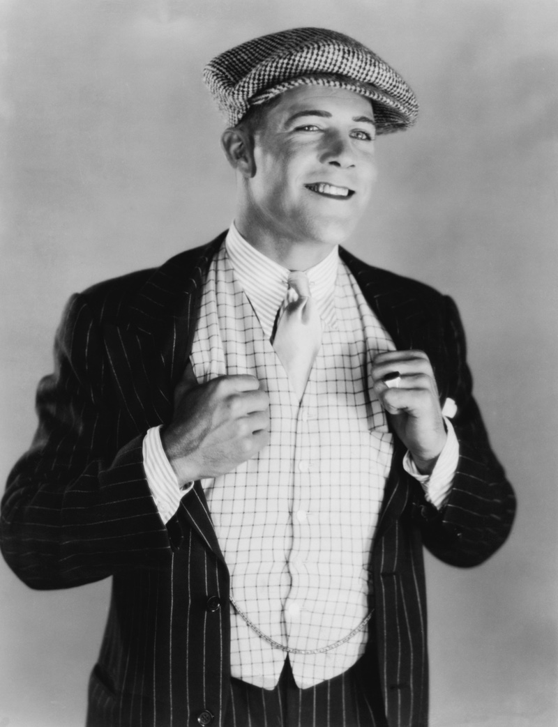 A 1920s man wears a stylish vest with a pinstripe suit, a striped shirt, and a collar pin along with a flat cap.