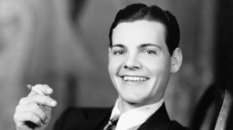 Flashing a smile, a young man in the 1920s wears a slick hairstyle featuring a side part.