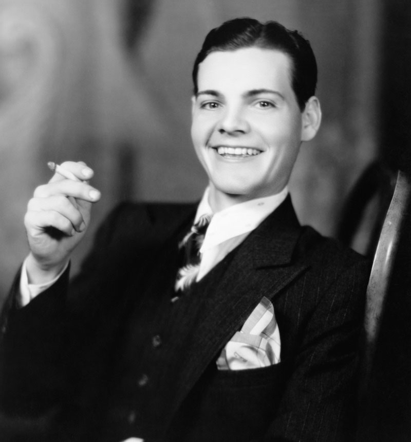 Flashing a smile, a young man in the 1920s wears a slick hairstyle featuring a side part.