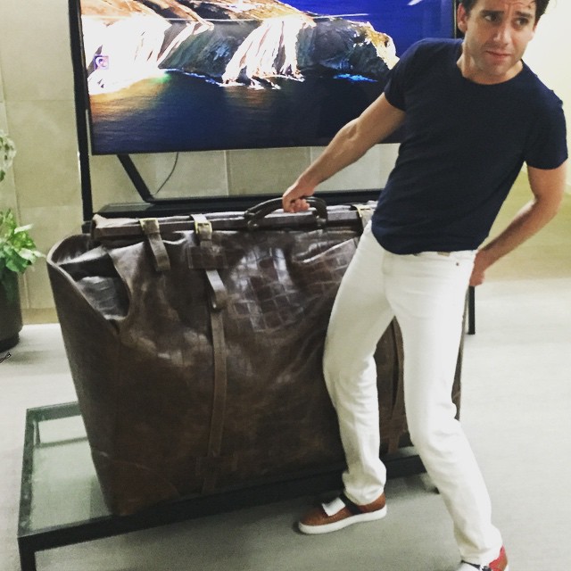 Sporting white denim jeans and a black tee, Mika poses for a cheeky image with oversized luggage.