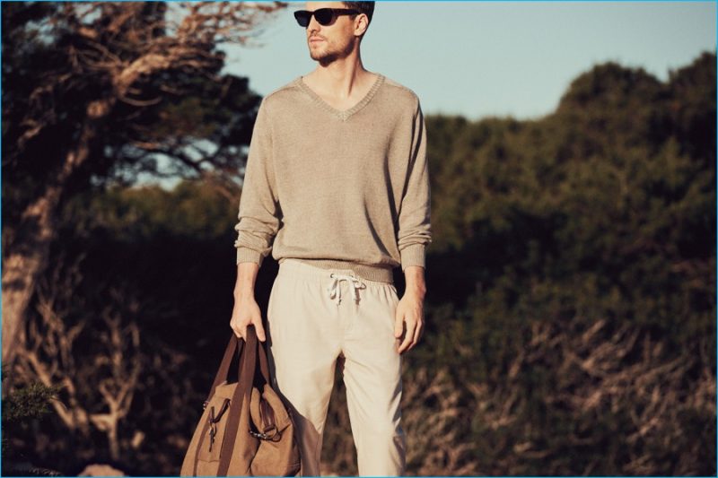 George Barnett models neutrals, wearing Mango Man's v-neck sweater and drawstring trousers, accessorized with a handbag and sunglasses.