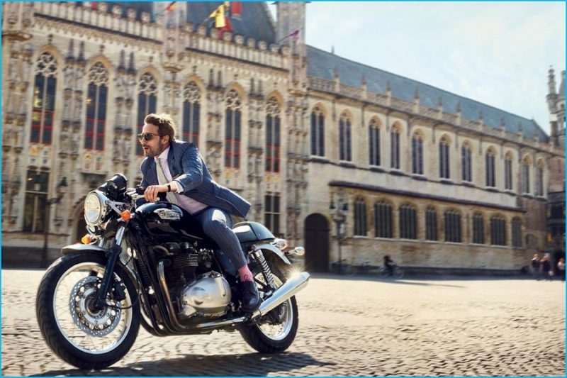 Matthias Schoenaerts gets on the back of a Triumph Bonneville 750 motorcycle, donning a pinstripe suit from Brunello Cucinelli with Garrett Leight sunglasses.