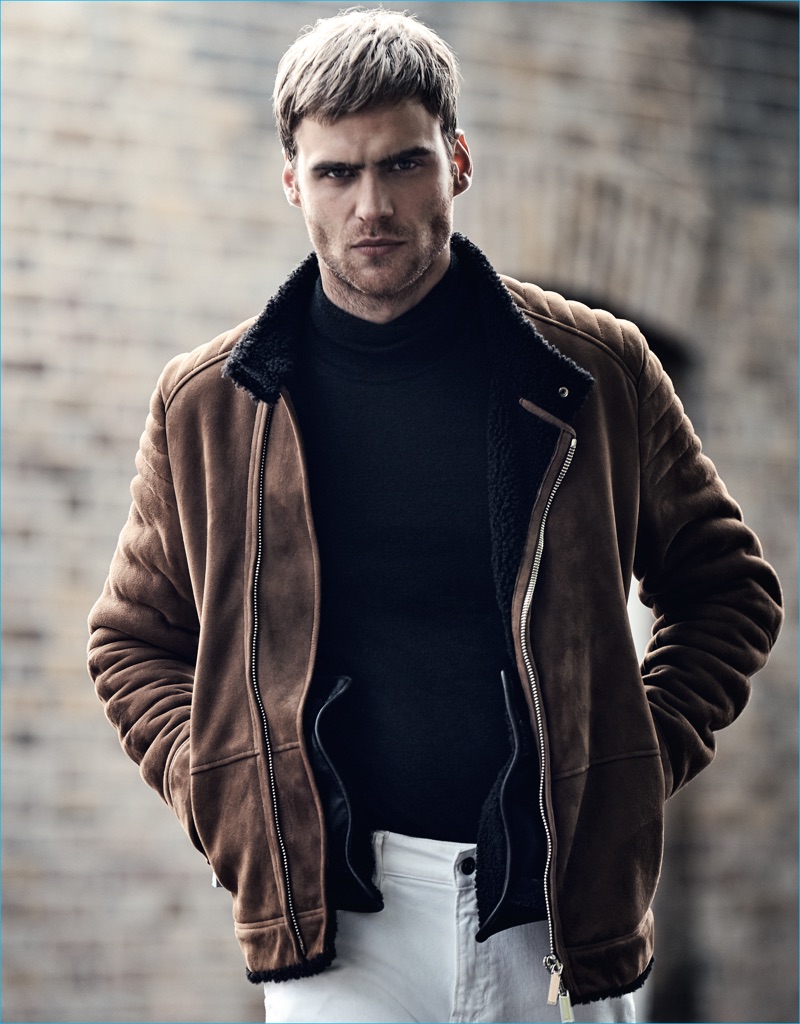 George Alsford dons a brown suede moto jacket with a mock turtleneck sweater, and white jeans from BOSS Hugo Boss for GQ France.