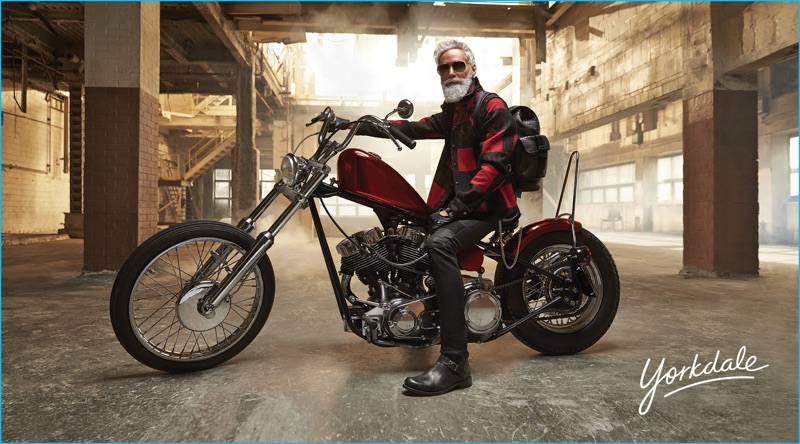 Fashion Santa wears a red and black Valentino check jacket with a Givenchy leather backpack. Sitting on the back of a motorcycle, Santa also sports an Acne Studios turtleneck, John Varvatos leather boots, Dents driving gloves, Christian Dior aviator sunglasses, and Belstaff leather pants.