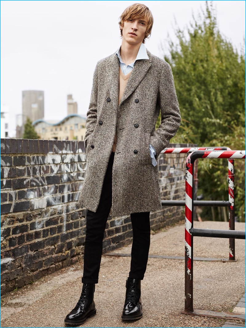 Tim Dibble dons a double-breasted herringbone coat with a cashmere sweater, striped shirt, black jeans, and boots by Zara Man.