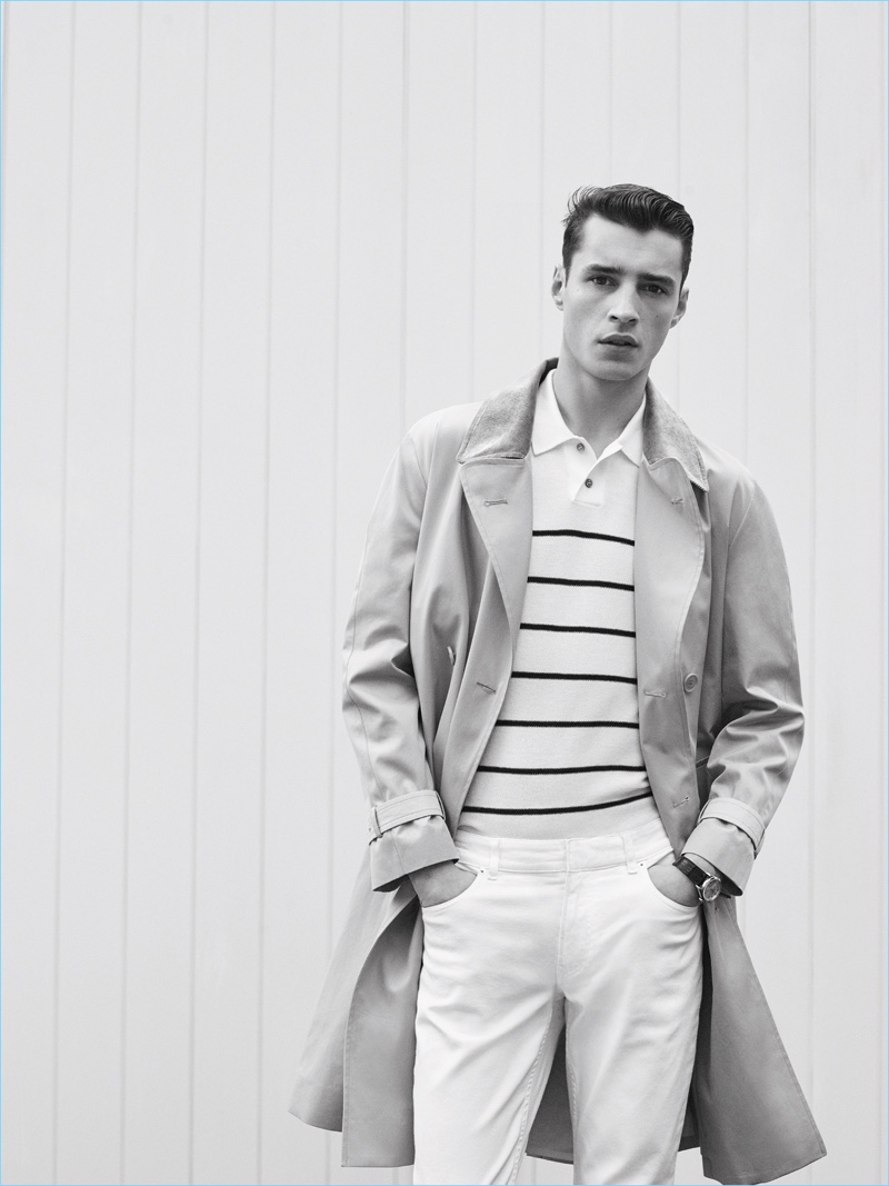 Stepping outdoors, Adrien Sahores wears a trench coat with a striped polo shirt and white pants by De Fursac.