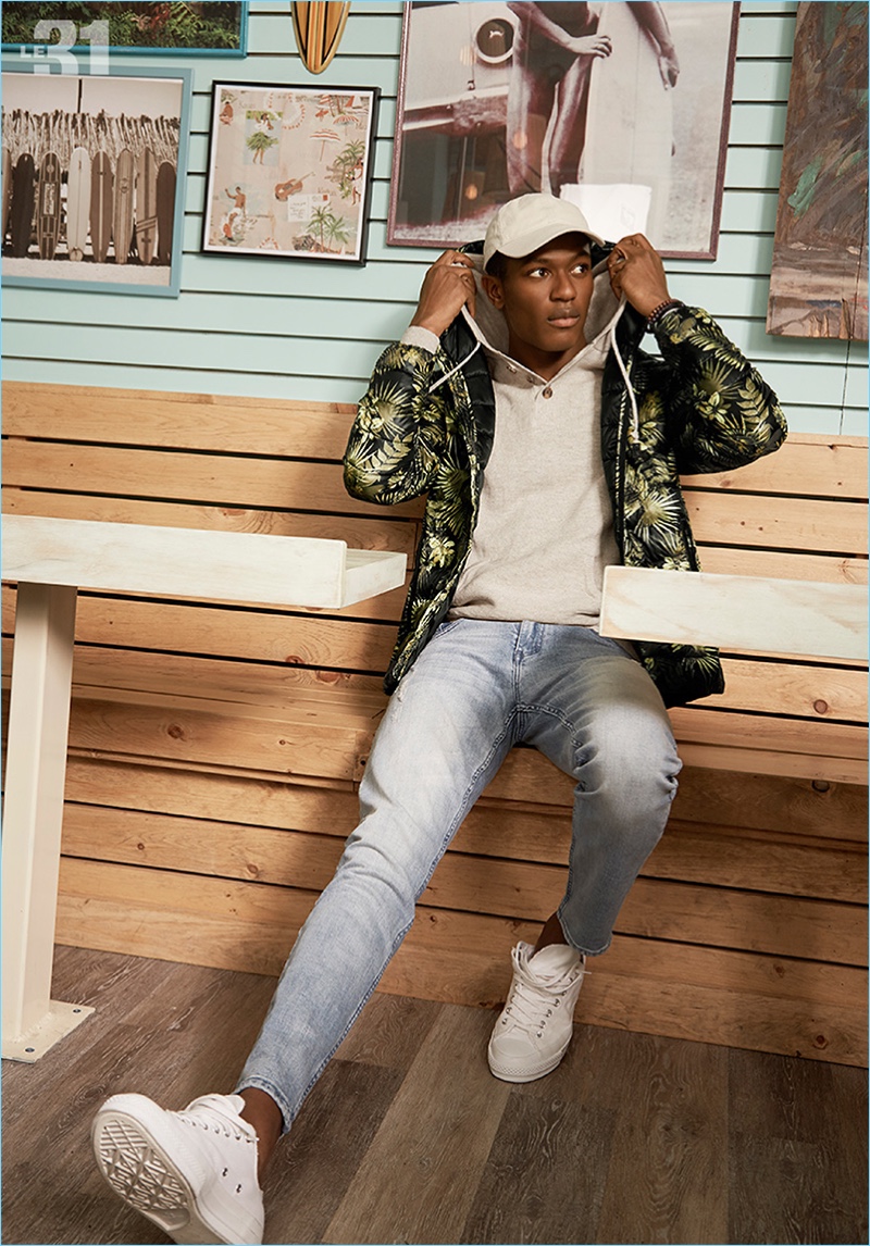 Tackling a sporty ensemble, Hamid Onifade models a LE 31 packable quilted jacket, henley style hoodie, white cap, and distressed denim jeans with Adidas Stan Smith sneakers.