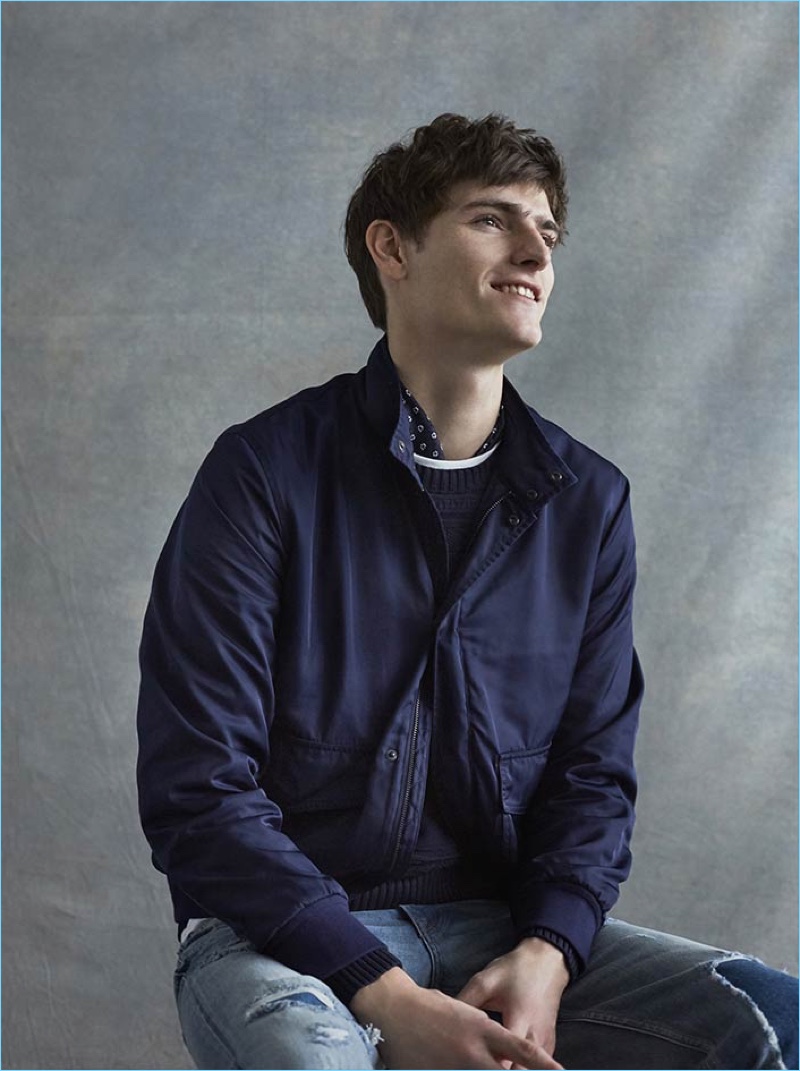 All smiles, Alexander Beck wears a technical newsboy jacket, ribbed sweater, floral scarf, and patchwork slim denim jeans from Club Monaco.
