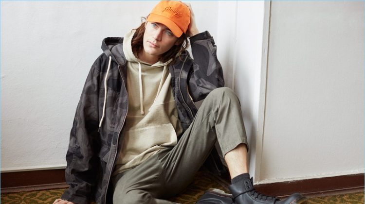 Model Jaco van den Hoven layers in a hoodie, camouflage print jacket, and twill pants with combat boots.