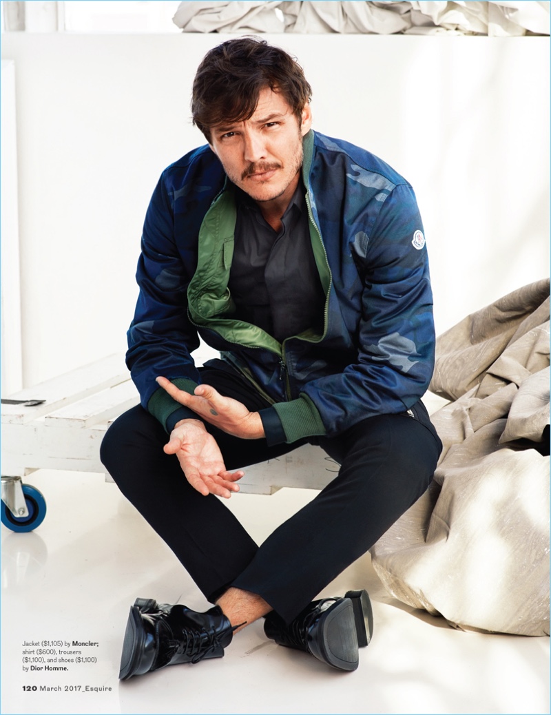 Pedro Pascal wears a camouflage jacket by Moncler with a shirt, trousers, and shoes by Dior Homme.