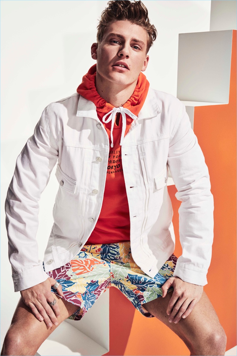 A colorful vision, Mikkel Jensen wears an orange hoodie with a white denim jacket and swim shorts by River Island.