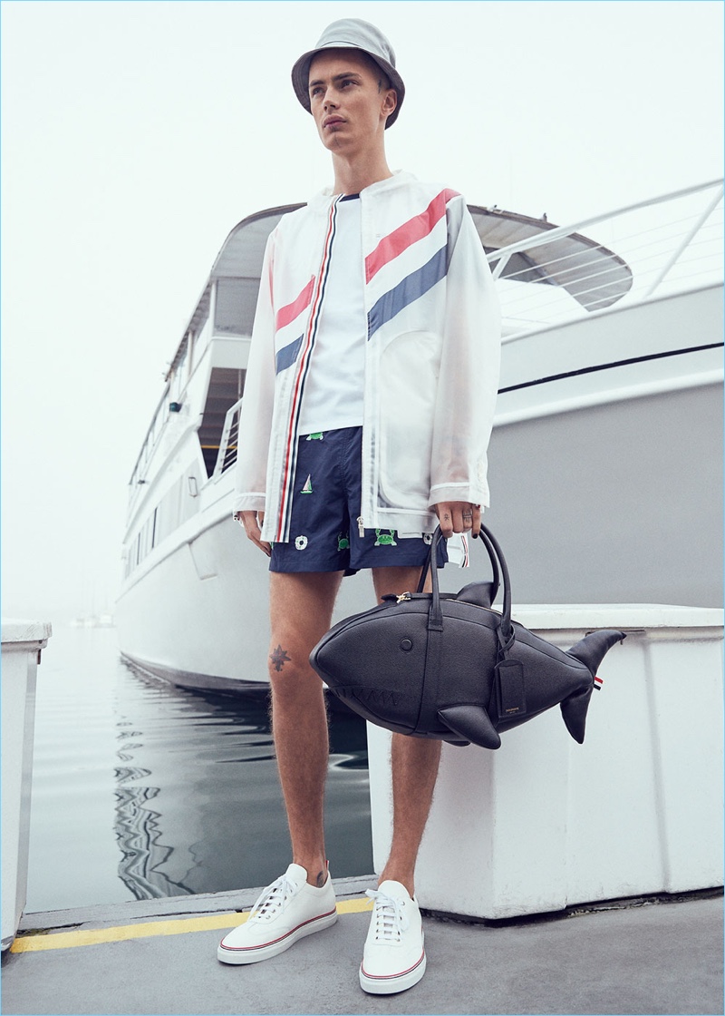 Model Simon Kotyk wears a Thom Browne diagonal stripe packable rain coat $2,490, fun mix jersey cotton short-sleeve tee $420, and embroidered swim trunks $412. The model also sports Thom Browne pebble grain trainers $590, a shark bag $2,500, and lined bucket hat $175 with Dries Van Noten gold sunglasses $345.