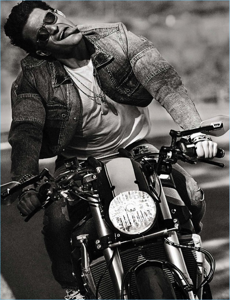 Riding a motorcycle, Orlando Bloom wears a t-shirt, denim jacket, and jeans by Valentino. Bloom also sports Visvim sneakers and Oliver Peoples sunglasses.