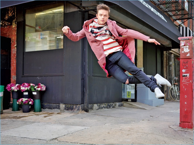 Leaping, Dane DeHaan wears a Stella McCartney t-shirt with a Prada overcoat. He also sports Saint Laurent jeans and Common Projects sneakers.