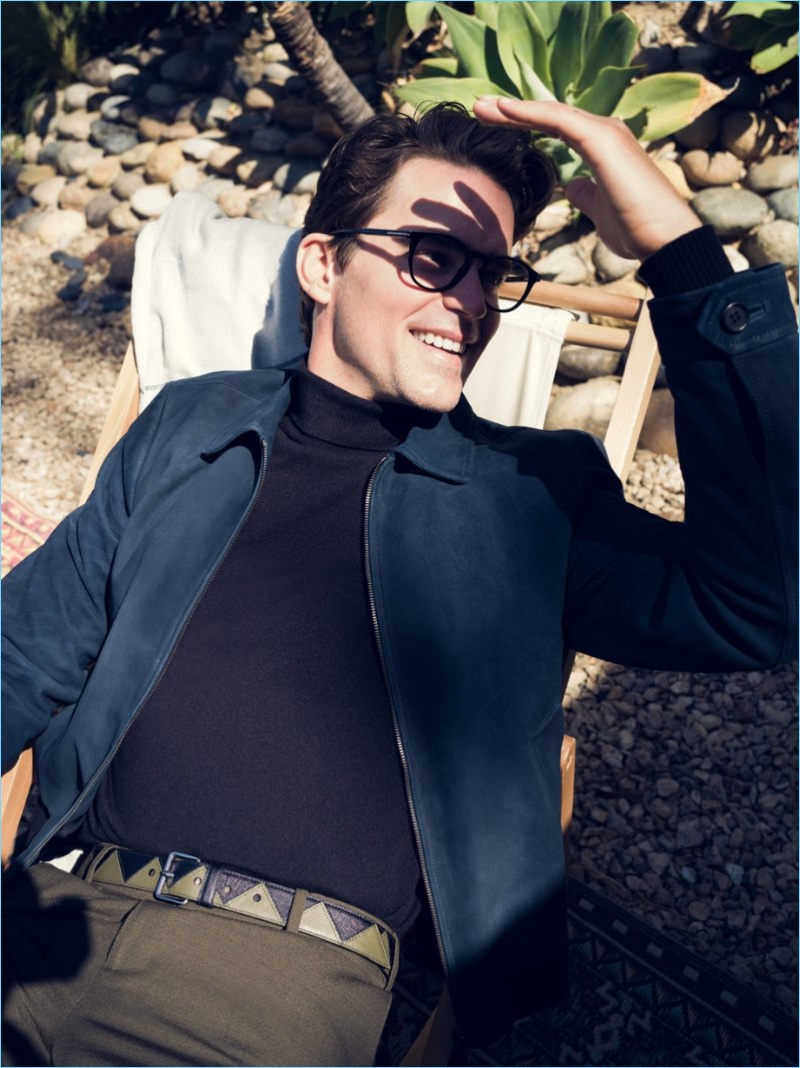 All smiles, Matt Bomer wears a Prada suede jacket, Maison Margiela turtleneck sweater, and Joseph trousers. Bomer adds some character to his look with a Prada two-tone leather belt and Tom Ford glasses.