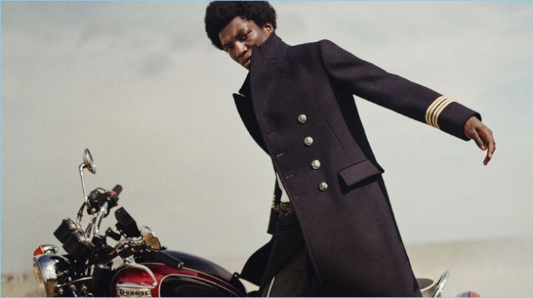 Model Josue Comoe connects with Farfetch for a fashion editorial. He wears a sharp officer coat by Gucci with an Alexander McQueen sweater. The model also sports John Lawrence Sullivan jeans with an Avant Toi scarf worn as a belt.