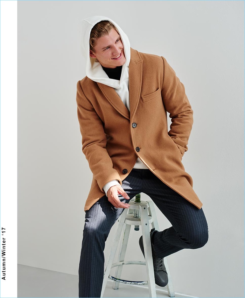 All smiles, Mikkel Jensen wears a camel single-breasted coat with pinstripe pants and a hoodie by Reserved.