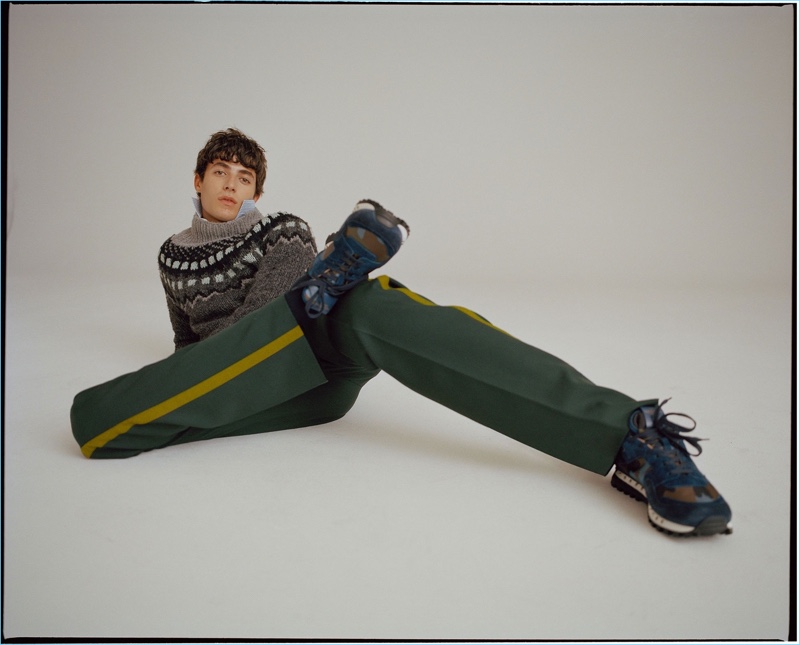 Relaxing, Oscar Kindelan wears a geometric sweater, striped shirt, trousers, and camouflage sneakers by Valentino.