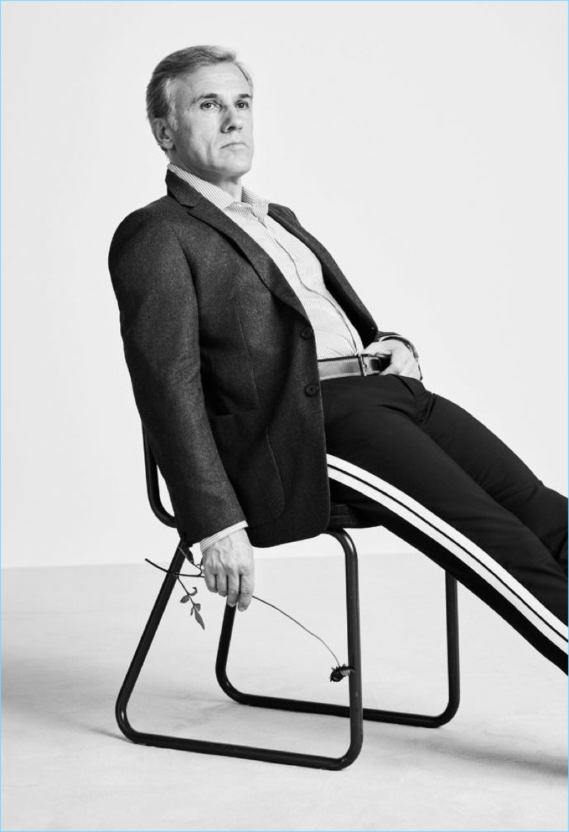 Sitting for a photo, Christoph Waltz sports a Prada blazer, Joseph shirt, Valentino trousers, and Loewe belt.