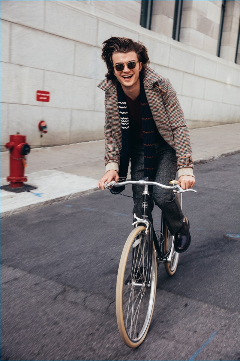 Taking a bike ride, Joe Keery wears a Prada jacket, sweater, scarf, and trousers. He accessorizes with Church's shoes and Steven Alan sunglasses.