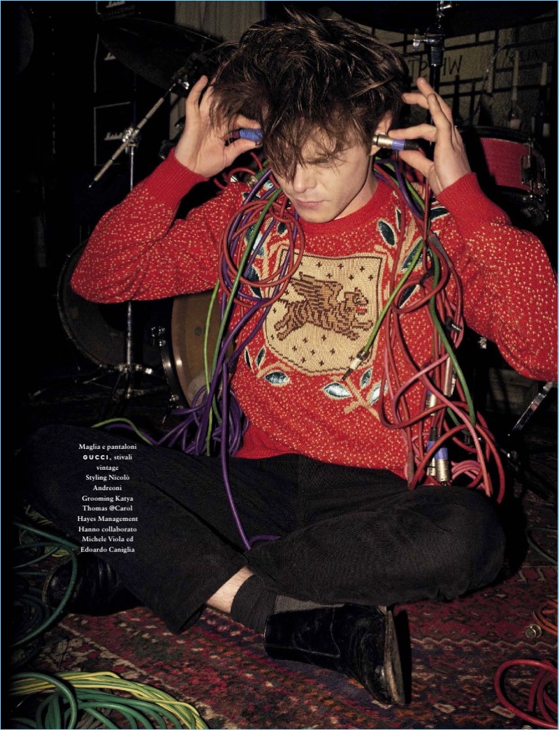 Appearing in a new photo shoot, Charlie Heaton wears a red Gucci sweater and black trousers.