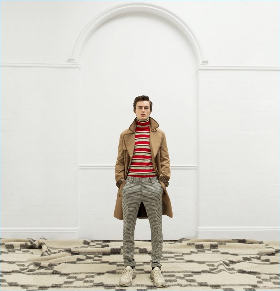 Jack Chambers models a double-breasted trench coat and tartan trousers by Fendi. The model also wears a striped Gucci sweater and leather sneakers.
