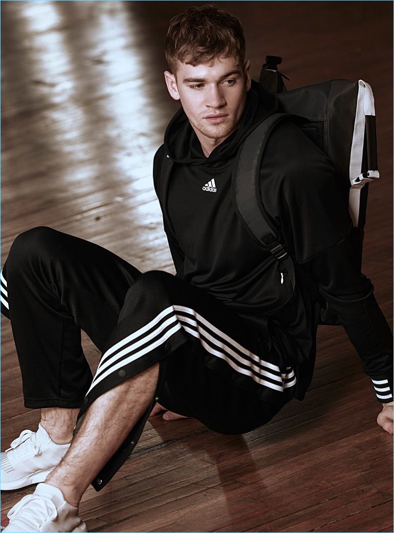 Relaxing, Matty Carrington wears an Adidas hoodie and snap button track pants with Adidas Originals sneakers.