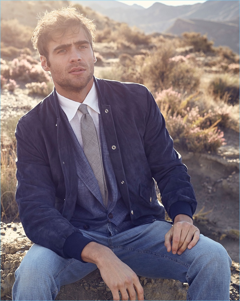 Sitting for a picture, George Alsford wears a Brunello Cucinelli suede bomber jacket in blue. He also models a wool tie, three-button pinstripe sport jacket, slim shirt, and jeans by Brunello Cucinelli.