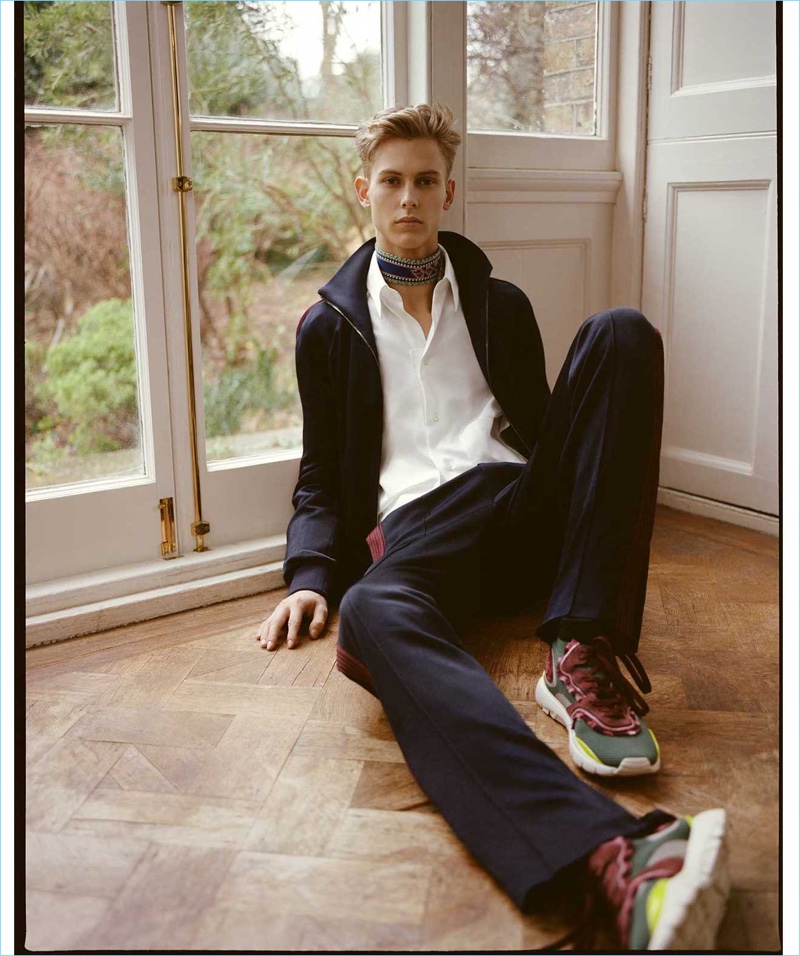 Connecting with Matches Fashion, Oliver Houlby wears a Valentino zip through track jacket and cotton-poplin shirt. The model also sports Valentino side-stripe track pants, Heroes high-top trainers and a multicolored logo-intarsia belt (worn as scarf).
