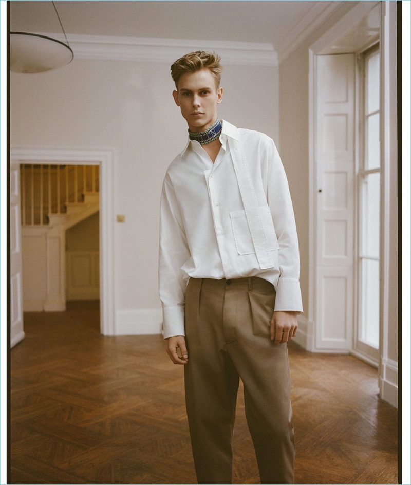 A smart vision, Oliver Houlby wears a strap-detail cotton-poplin shirt, logo-print chinos, and a multicolored logo-intarsia belt (worn as scarf) by Valentino.