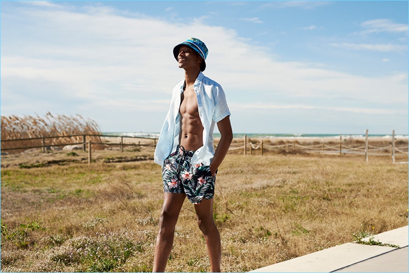 All smiles, Oliver Kumbi wears bucket hat, open shirt, and tropical print swim shorts by Tommy Hilfiger.