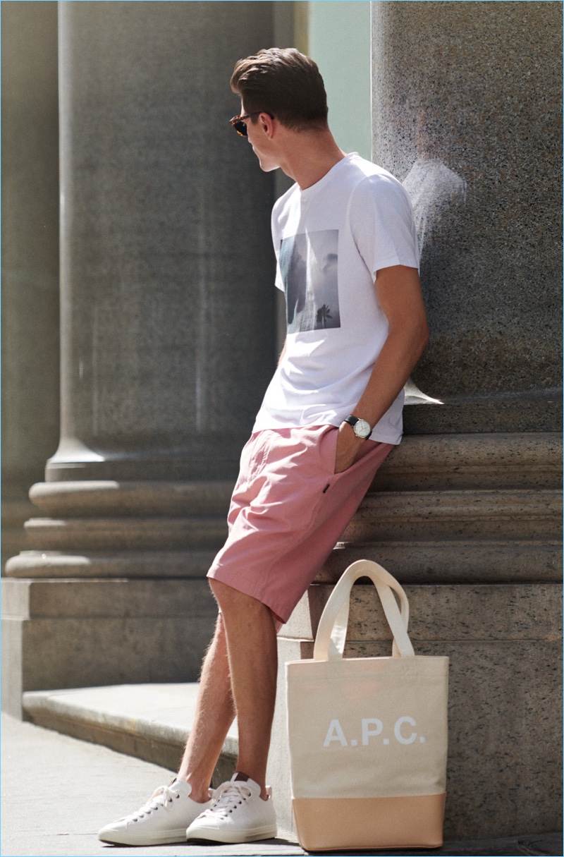 Eli Hall wears an A.P.C. graphic tee with Stüssy shorts, a Larsson & Jennings Meridian watch, and Coach New York sneakers. He accessorizes with an A.P.C. tote and Oliver Peoples sunglasses.