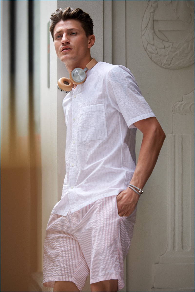 Stepping out in style, Eli Hall models a Theory pinstripe shirt with Obey shorts. The model also sports a Miansai wrap bracelet and Bang & Olufsen H8i wireless headphones.