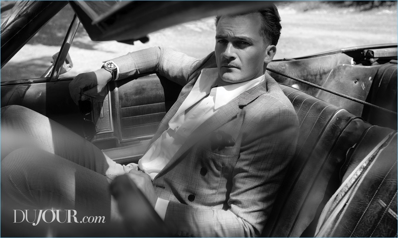Sitting in a vintage car, Rupert Friend wears a Bottega Veneta suit with a Brioni shirt, Dolce & Gabbana pocket square, and Breitling watch.