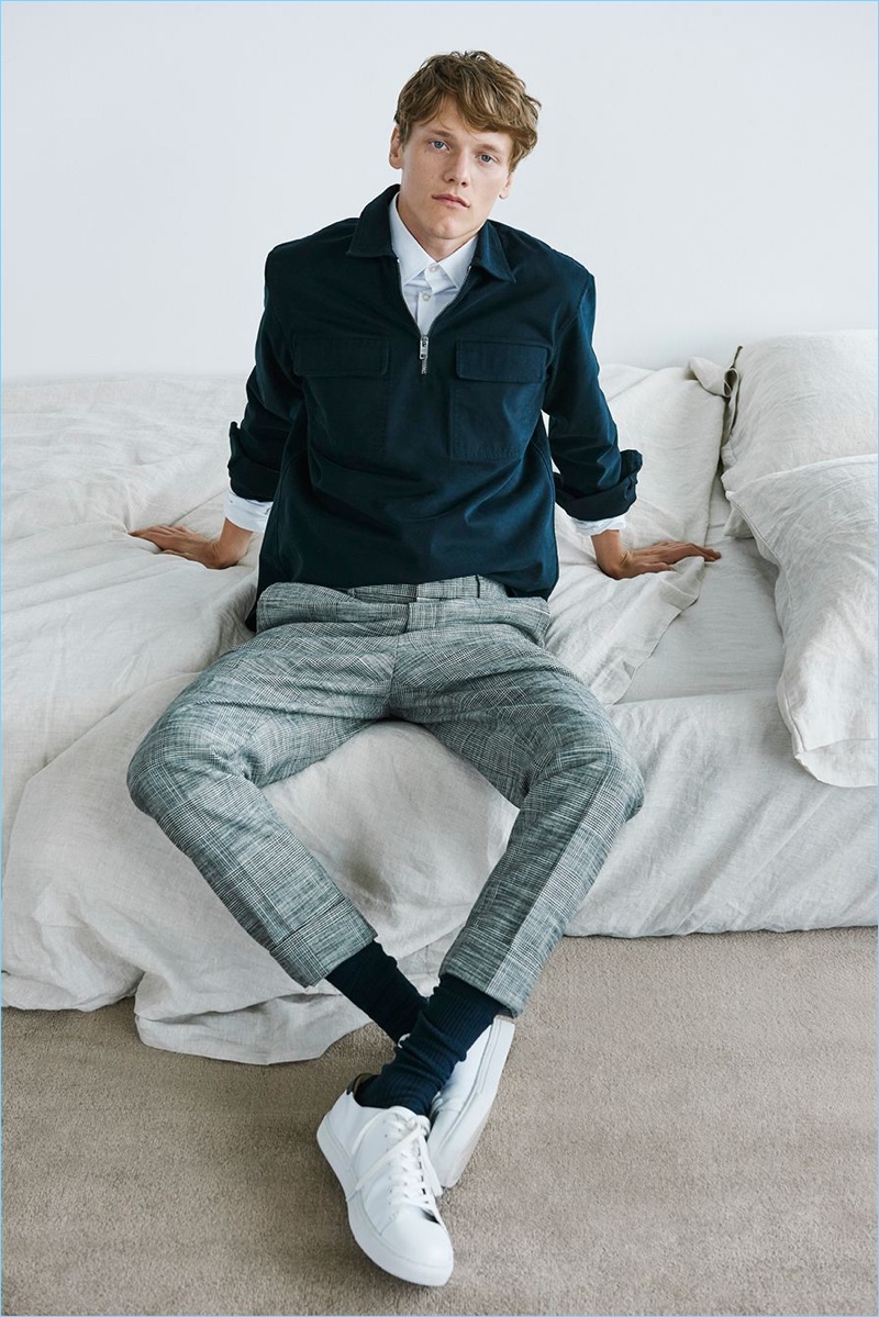 Ryan Keating dons a dress shirt, zip work shirt, pants, and white sneakers by Club Monaco. 