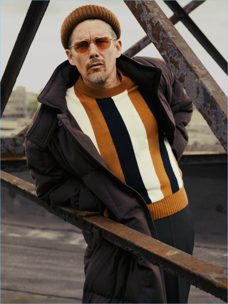 A cool vision, Ethan Hawke wears a BOSS coat, pants, and beanie. Hawke also rocks a Sandro sweater and Garrett Leight sunglasses.
