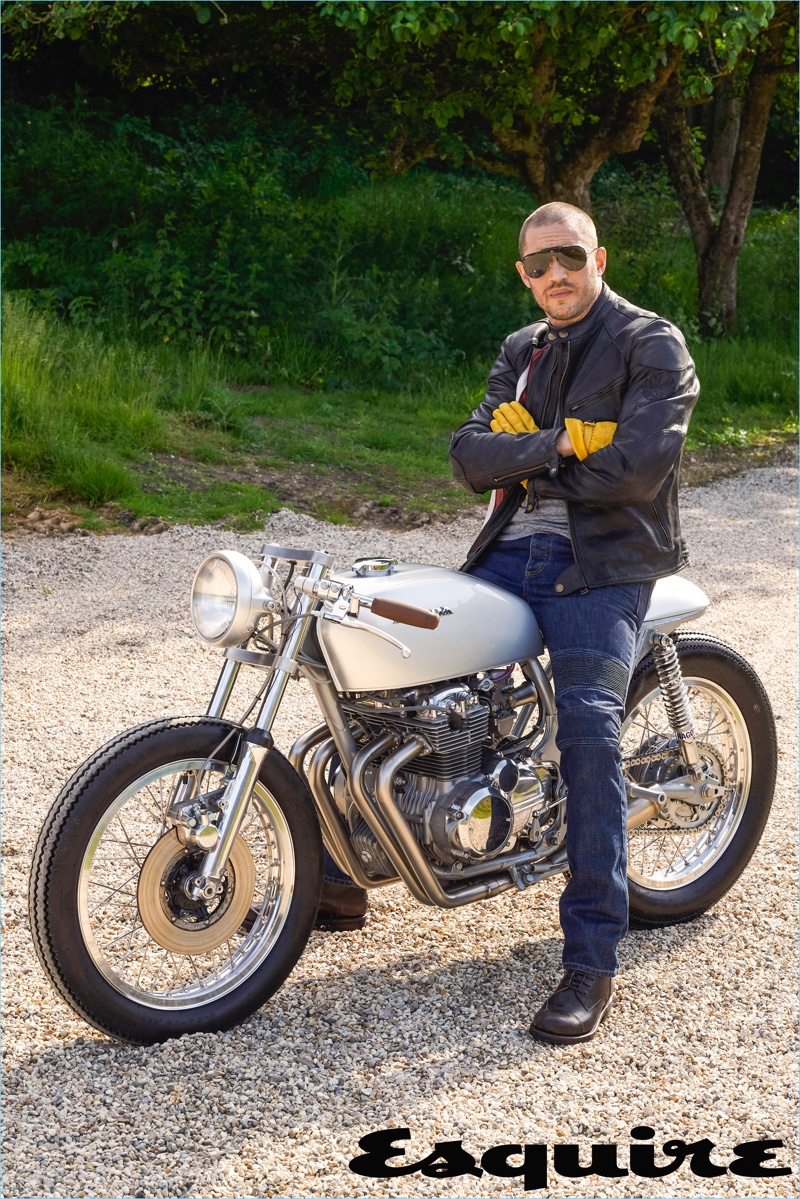 Sitting on a bike, Tom Hardy wears a leather jacket and jeans by Belstaff. He also sports an Alternative Apparel t-shirt, Triumph boots, and Carrera sunglasses.