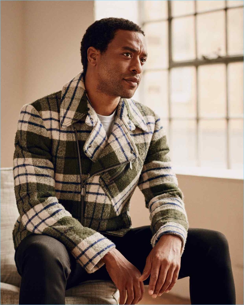 Sitting for a photo, Chiwetel Ejiofor dons a Bottega Veneta biker jacket, Brunello Cucinelli t-shirt, and Acne Studios jeans.