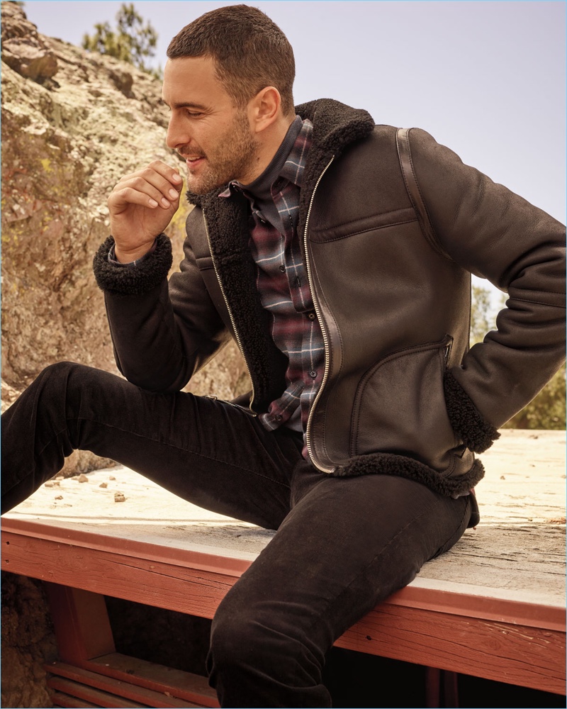 Posing outdoors, Noah Mills wears a Vince shearling jacket, plaid shirt, corduroy pants, and a turtleneck sweater.